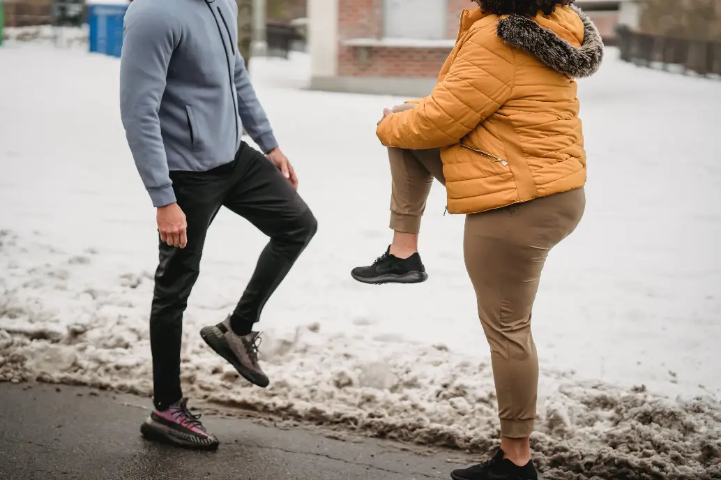 Side view of unrecognizable man and woman with raised legs warming up while preparing for training in winter park on street
